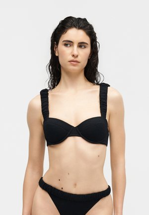 Γυναίκα με βρεγμένα σκούρα μαλλιά που φοράει μαύρο μπικίνι με υφή και ραβδώσεις στις τιράντες, κοιτάζοντας ελαφρώς προς τα αριστερά της, με απλό ανοιχτόχρωμο φόντο.