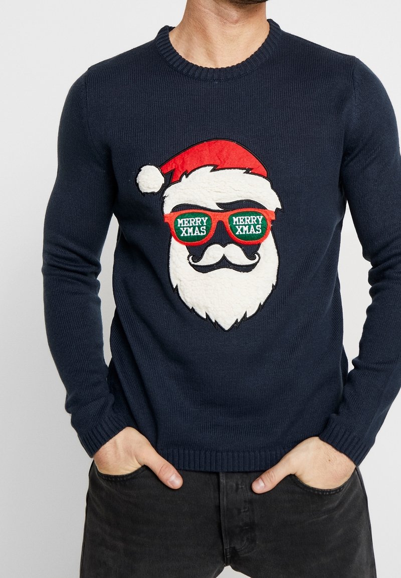 Pull marine avec un visage de Père Noël texturé portant un chapeau rouge et des lunettes de soleil rouges avec l'inscription "MERRY XMAS", porté par une personne les mains dans les poches.