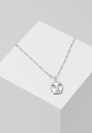 Collier en chaîne argentée avec un pendentif en forme de visage abstrait, présentant un nez en relief et des zones des yeux texturées, présenté sur un bloc géométrique blanc.