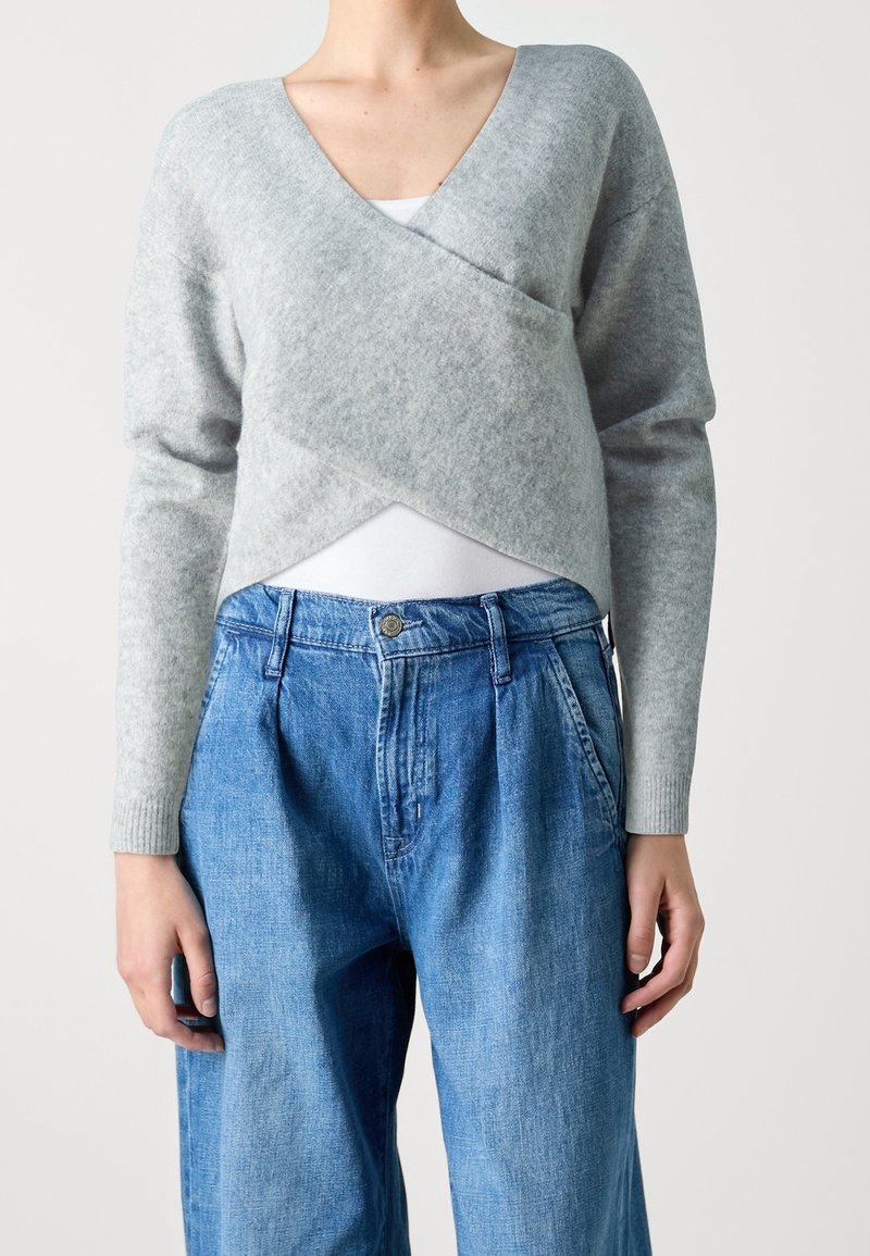 Femme portant un pull cache-cœur gris clair par-dessus un haut blanc et un jean bleu ample taille haute, debout devant un fond uni.