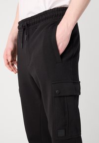 Gros plan sur un pantalon cargo noir avec ceinture élastique, poches latérales, poche à rabat avec boutons, et une main placée dans l'une des poches.