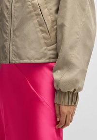 Beige bomber jacket with a textured finish, featuring a zip closure and ribbed cuffs, paired with a bright pink satin skirt.