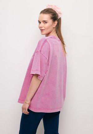 Młoda kobieta z długimi włosami związanymi różową gumką, ubrana w luźny różowy T-shirt i ciemne dżinsy, patrząca przez ramię.