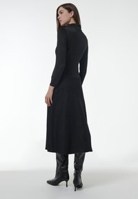 Zwart langemouwen top gecombineerd met een donkergrijze rok tot de knieën. Model draagt kniehoog zwarte laarzen. Gladde textuur met een aansluitende snit.