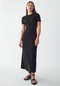 Schwarzes, strukturiertes Midi-Kleid mit kurzen Ärmeln, tailliertem Oberteil und weitem Saum, kombiniert mit schwarzen Riemchensandalen. Glatte Stoffoberfläche.