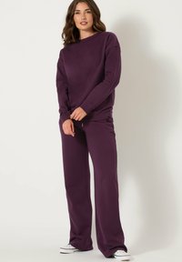 Nainen seisoo päällään matching tumman violetti huppari ja leveälahkeiset housut, valkoiset lenkkarit jalassa, yksinkertaista vaaleaa taustaa vasten.