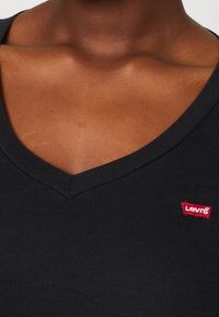 Černý žebrovaný top s výstřihem do V a červenou visačkou Levi's na levé straně. Výstřih je dobře definovaný a materiál vypadá měkce.