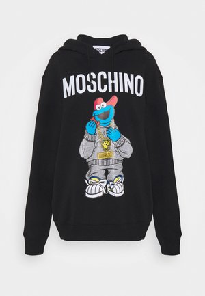 Henkilö, jolla on päällään musta Moschino-huppari Cookie Monster -kuvalla, mustavalkoraidalliset housut, mustat niittisaappaat ja kädessään punainen olkalaukku.