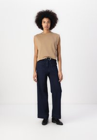 Top bege sem mangas combinado com jeans de perna larga azul escuro com bolsos frontais e um cinto preto. Botas são pretas com um salto baixo.