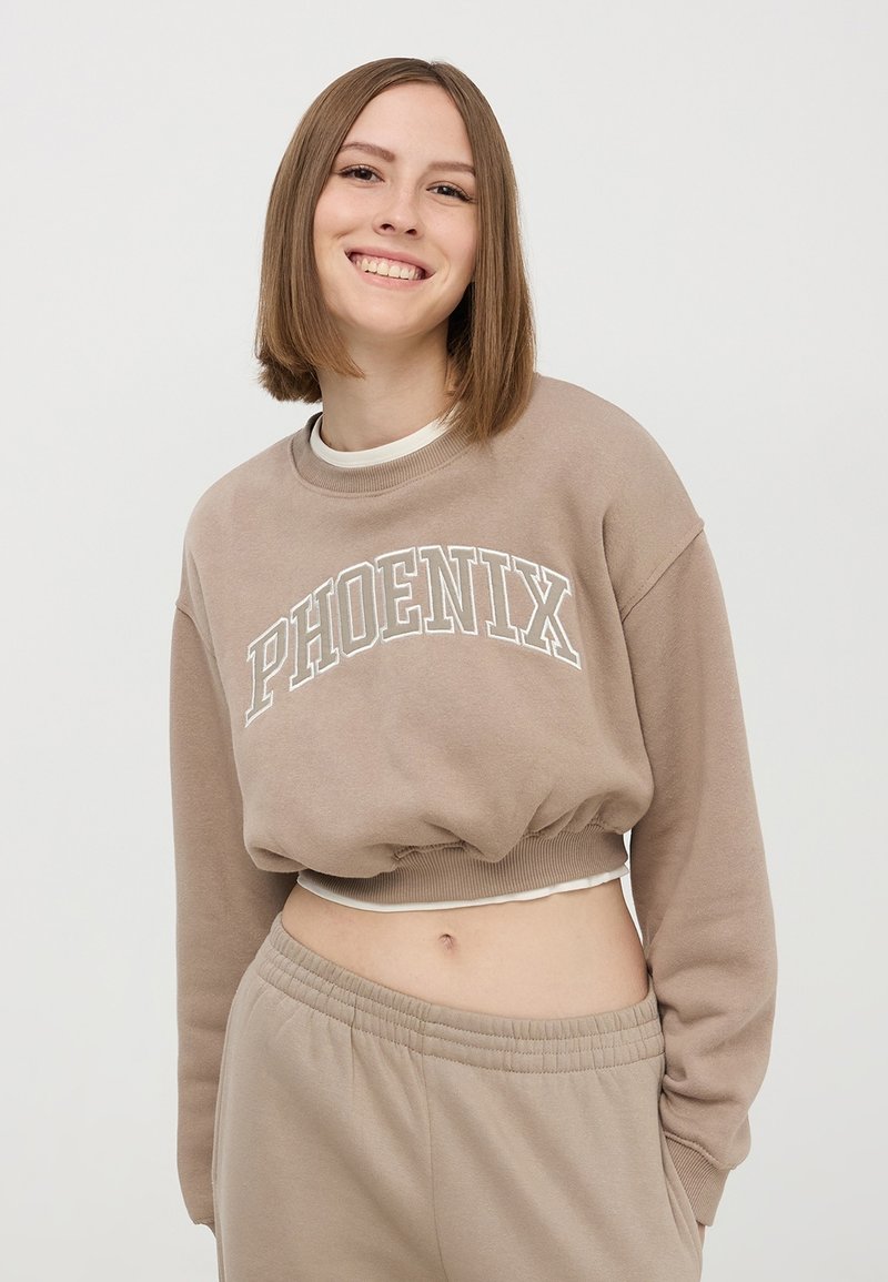 Beżowa krótka bluza z nadrukiem "PHOENIX", z ryflowanym dekoltem i wykończeniem, zestawiona z dopasowanymi dresami, ukazująca miękki materiał i luźny krój.
