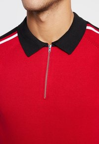 Polo rouge avec un col noir, doté d'une fermeture éclair moitié devant et de bandes blanches le long des épaules. Tissu texturé.