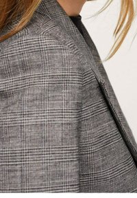 Szara marynarka w kratę wykonana z fakturowanej tkaniny, z usztywnionymi ramionami i gładkim zamkiem błyskawicznym z przodu, prezentująca nowoczesny design.