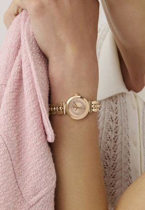 Goudkleurig polshorloge met een ronde wijzerplaat, versierd met kristallen, bevestigd aan een schakelarmband. Het horloge wordt gedragen aan een arm die een roze textuurtrui vasthoudt.