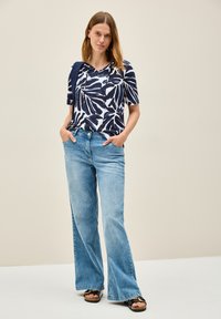 Camisa de flores en tonos marino y blanco, de manga corta, con capucha, combinada con jeans de mezclilla de pierna amplia en azul claro y sandalias negras.