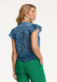 Zila un zaļa paisley raksta blūze ar Bura piedurknēm, izgatavota no viegla auduma; savienota ar zaļām biksēm. Redzami zeltaini riņķa auskari.