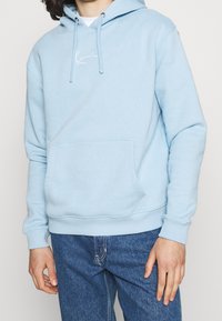 Light blue hoodie made of cotton blend fabric. Features a front kangaroo pocket, drawstring hood, and white embroidered logo on chest.
