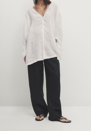 Woman wearing loose white long-sleeve blouse, wide black pants, and black flip-flops standing against a plain light background.