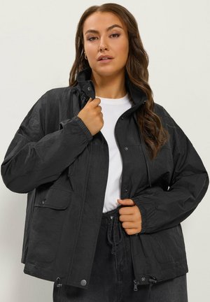 Femme ajustant le col d'une veste noire ample sur une chemise blanche, avec de longs cheveux ondulés, debout devant un fond clair uni.