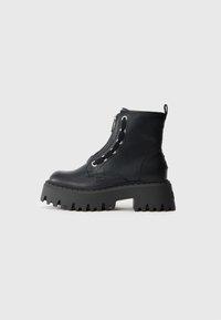 Bota de cuero negro hasta el tobillo con suela gruesa de tracción, cremallera frontal, ojales para cordones y etiqueta de marca en el costado.