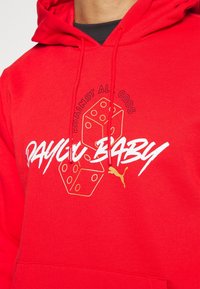 Sweat à capuche rouge avec le texte blanc "DAYGU BABY", des dés entourés d'or, "AGAINST ALL ODDS" au-dessus des dés, et un petit logo Puma doré.