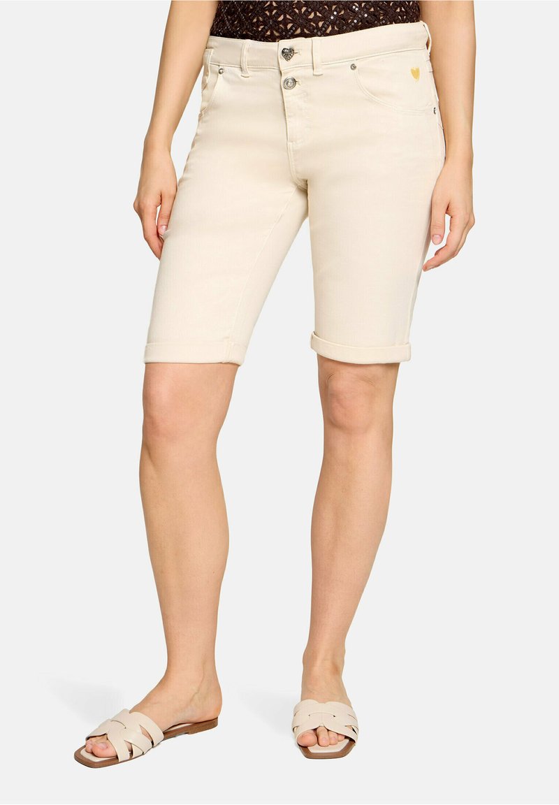 Femme portant un short crème mi-long avec ourlet roulé, un top en dentelle noire et des sandales beige à bout ouvert, debout sur un fond uni.