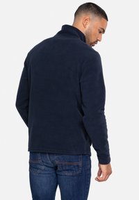 Pullover in pile blu navy con colletto alto, cuciture piatte e maniche lunghe; abbinato a jeans in denim, che mostrano un tessuto morbido e testurizzato.