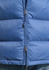 Modrá zateplená vesta s hladkým povrchem, která má dvojitý zip, obdélníkové prošívané sekce a nastavitelné stahování v límci.