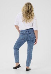 Jeans de mezclilla azul de talle alto con un lavado desgastado, que cuentan con bolsillos traseros y un corte ajustado hasta el tobillo, combinados con una camisa blanca de manga corta.