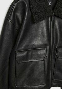 Czarna skórzana kurtka z teksturowanym czarnym kołnierzem z owczej skóry, przednim zamkiem błyskawicznym i dużymi klapami kieszeni po obu stronach.