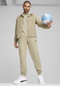 Beige træningssæt bestående af en knap-op jakke og matchende bukser, lavet af blødt stof. Inkluderer hvide sneakers med sort accent og en blå fodbold.