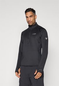 Man draagt een zwart Columbia Titanium long-sleeve pullover met een kwart rits en duimhole, staande tegen een effen achtergrond.