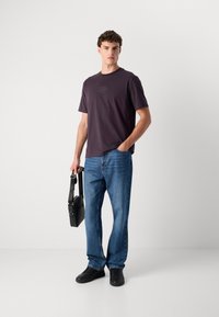 Szürke póló kis logóval, kék egyenes lábszárú farmerrel párosítva. Férfi, aki egy kis fekete keresztpántos táskát tart, és fekete sportcipőt visel.