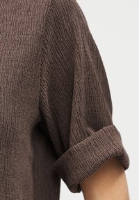 Καφέ υφασμάτινο κοντομάνικο πουκάμισο με γυρισμένο μανίκι στο χέρι ενός ατόμου, πάνω σε απλό φωτεινό φόντο.