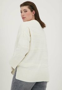 Pulover de crem deschis cu un model texturat de tricot, margine côtelată și mâneci până la trei sferturi, purtat peste blugi gri închis.