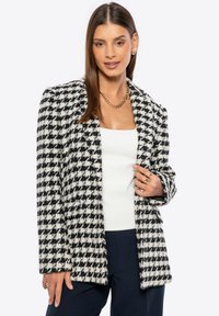 Blázer de pata de gallo en blanco y negro con una textura suave, solapas en pico y bolsillos frontales, usado sobre una blusa blanca y pantalones oscuros.