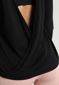 Schwarzes langärmliges Oberteil mit einem tiefen Rückenausschnitt, ausgestattet mit drapiertem Stoff und glatter Textur. Kombiniert mit hellrosa, enganliegenden Leggings.