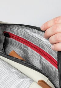 Innenansicht einer Tasche mit einem grauen Karomuster-Futter, einer roten Reißverschlusstasche und einem beige Lederakzent.