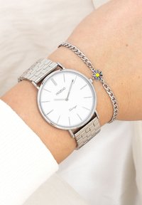 Montre en acier inoxydable avec un bracelet en maille argentée et un cadran blanc. Présente des index heures fins, ainsi qu'un délicat bracelet en chaîne argentée avec un charm floral.