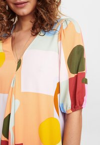 Robe colorée avec un décolleté en V, des manches courtes bouffantes et un motif abstrait de formes dans des tons pastel et terre. Texture de tissu douce.