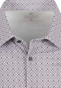 Witte blouse met een geometrisch patroon in rood, marineblauw en grijs. Beschikt over een klassieke kraag en een enkele grijze knop bij de halsopening.