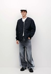 Quiltad marinblå jacka med rund halsringning och två framfickor, i kombination med slitna grå jeans med vida ben och svarta klumpiga skor.