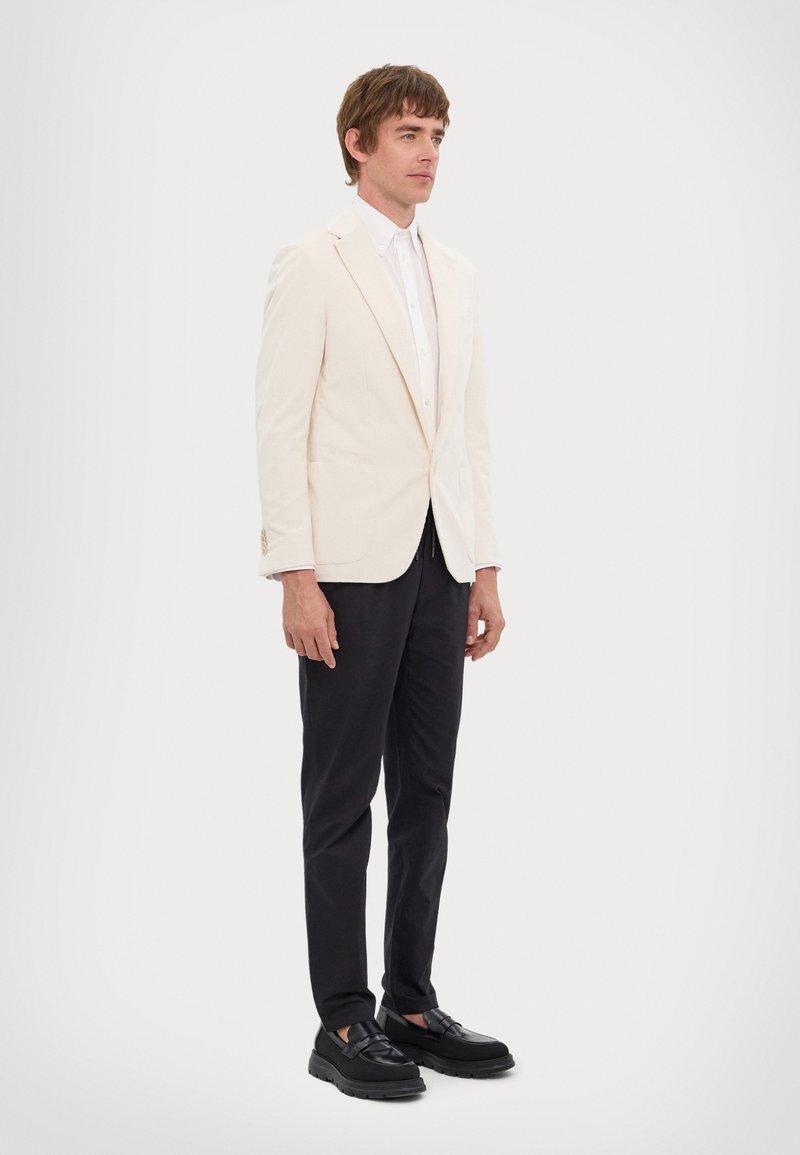 Hele beež blazer tekstureeritud kangast, valge särk all, mustad püksid ja mustad paksu tallaga kingad, seismas ühtlases taustas.