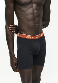 Μυώδης άτομο που φοράει μαύρα μποξεράκια Nike με πορτοκαλί ζώνη στη μέση που φέρει λογότυπα Nike και σύμβολα swoosh.
