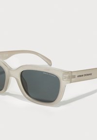 Permatomi smėlio spalvos akiniai nuo saulės su tamsiais lęšiais, šiek tiek suapvalinta stačiakampio forma ir "ARMANI EXCHANGE" ženklinimas ant šventės.