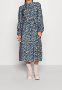 Floral dress in blue and green tones with long sleeves, a buttoned front, and a waist tie. Paired with light pink, chunky ankle boots.
