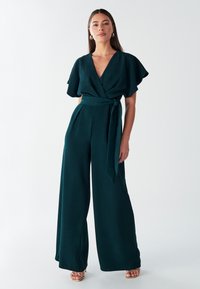 Tumši zaļš platajām kājām kombinezons ar īsām plīvura piedurknēm, V veida izgriezumu un jostu viduklī. Gluda, bezrakstu auduma.