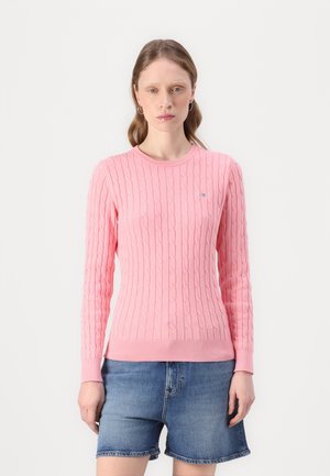 Rosa gestrickter Pullover mit Kabelmuster und geripptem Saum. Lange Ärmeln und Rundhalsausschnitt. Kombiniert mit Denim-Shorts. Kleines Logodetail auf der Brust.