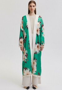 Pikk, roheline kimono lillemustriga, valge viimistlus ja tutid varrukatel, koos neutraalse ülaosa ja heledate pükstega.