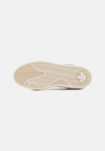 Σόλα από μπεζ λαστιχένιο αθλητικό παπούτσι με ζιγκ ζαγκ σχέδιο και ανάγλυφο λογότυπο φύλλου σφενδάμου κοντά στη φτέρνα, σε απλό λευκό φόντο.