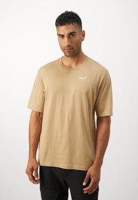T-shirt in cotone beige con collo rotondo e maniche corte, caratterizzato da un piccolo logo bianco nella zona sinistra del petto. Texture liscia.